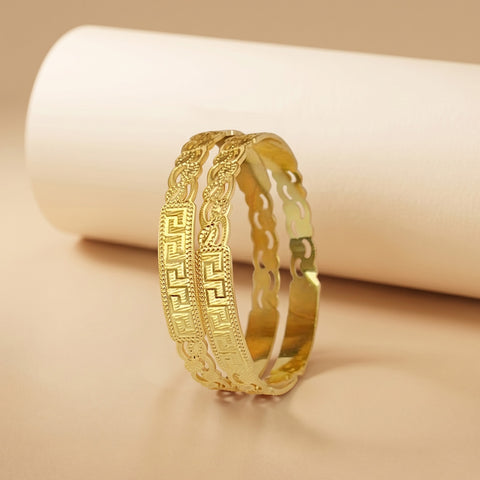 A pair of gold-plated bangles with intricate Greek key and floral engravings, displayed on a soft beige surface with a white cylindrical prop in the background.