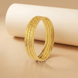 A textured gold bangle bracelet with multiple parallel ridges, displayed on a warm beige surface with a white cylindrical prop in the background.