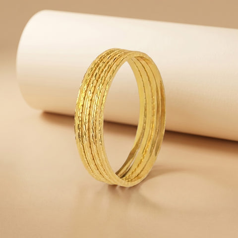 A textured gold bangle bracelet with multiple parallel ridges, displayed on a warm beige surface with a white cylindrical prop in the background.