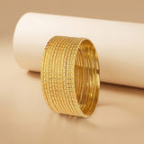 A stack of gold bangles with textured, dotted patterns, displayed on a warm beige surface in front of a white cylindrical prop.