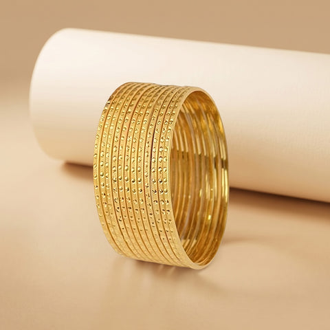 A stack of gold bangles with textured, dotted patterns, displayed on a warm beige surface in front of a white cylindrical prop.