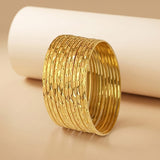 A stack of wide gold bangles with textured, diamond-patterned outer surfaces and smooth inner bands, displayed on a warm beige surface with a white cylindrical prop in the background.