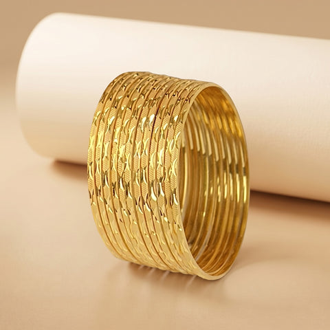 A stack of wide gold bangles with textured, diamond-patterned outer surfaces and smooth inner bands, displayed on a warm beige surface with a white cylindrical prop in the background.