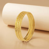 A gold-toned textured bangle bracelet standing upright in front of a white cylindrical prop on a warm beige background.