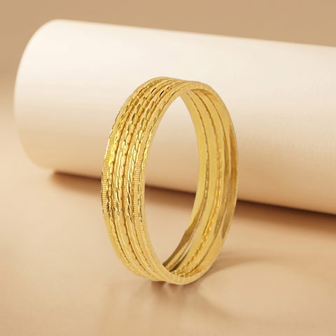 A gold-toned textured bangle bracelet standing upright in front of a white cylindrical prop on a warm beige background.