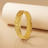 A gold bangle bracelet with a textured, woven leaf-like pattern on the outer band, displayed on a warm beige surface with a white cylindrical prop in the background.