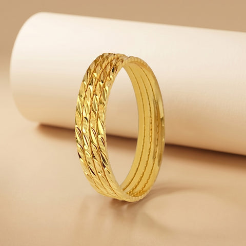 A gold bangle bracelet with a textured, woven leaf-like pattern on the outer band, displayed on a warm beige surface with a white cylindrical prop in the background.