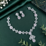 Silver-toned jewelry set with a necklace, earrings, and bracelet featuring geometric diamond motifs, floral links, and teardrop-shaped pink stones, arranged on a dark green satin fabric background with decorative greenery and flowers.