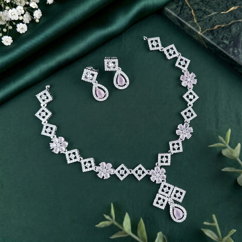 Silver-toned jewelry set with a necklace, earrings, and bracelet featuring geometric diamond motifs, floral links, and teardrop-shaped pink stones, arranged on a dark green satin fabric background with decorative greenery and flowers.