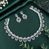Silver-toned jewelry set featuring a necklace and matching earrings with ornate floral motifs set with pink gemstones, displayed on a dark green satin fabric background with decorative green leaves and white flowers.