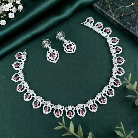 Silver-toned jewelry set featuring a necklace and matching earrings with ornate floral motifs set with pink gemstones, displayed on a dark green satin fabric background with decorative green leaves and white flowers.