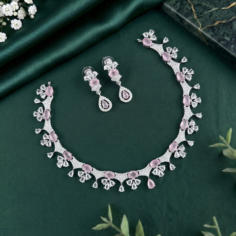 A silver-toned jewelry set featuring a necklace and matching drop earrings with pink gemstone accents, arranged on a dark green satin fabric background with decorative floral props.
