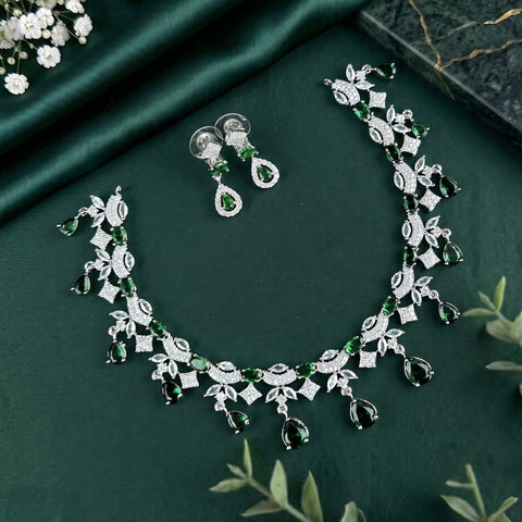 A silver-toned jewelry set featuring a necklace and matching earrings, decorated with green teardrop gemstones and clear pavé-style stones, arranged on a dark green satin fabric background with decorative floral props.