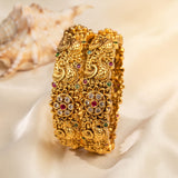 Two ornate gold-toned bangles with intricate filigree detailing and small red and green stone accents, displayed on draped cream satin fabric with a seashell in the background.