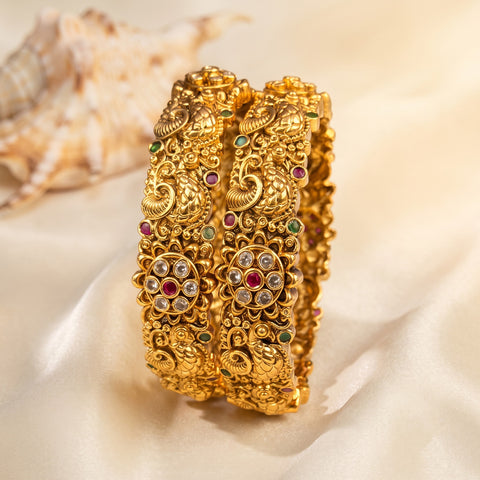 Two ornate gold-toned bangles with intricate filigree detailing and small red and green stone accents, displayed on draped cream satin fabric with a seashell in the background.