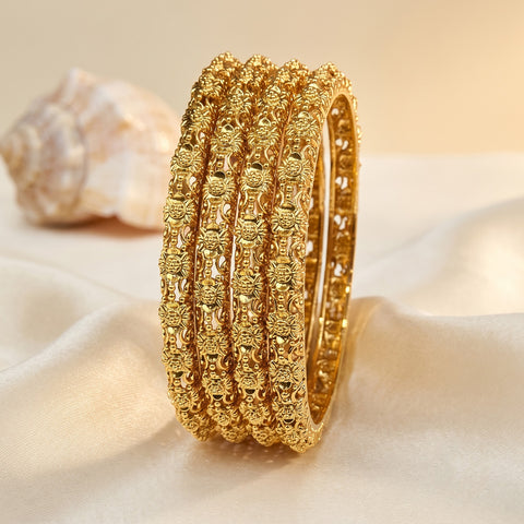 A stack of five textured gold bangles displayed on a soft cream satin surface with a decorative seashell in the background.