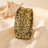 An ornate gold-toned bangle bracelet with intricate filigree detailing and small embedded red, green, and white stones, displayed on a beige satin surface with a seashell in the background.