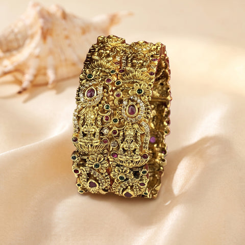 An ornate gold-toned bangle bracelet with intricate filigree detailing and small embedded red, green, and white stones, displayed on a beige satin surface with a seashell in the background.