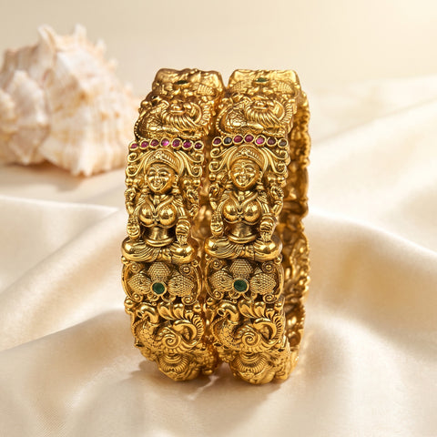 A pair of ornate gold-toned bangles with detailed deity carvings and small colored gem accents, displayed on draped cream satin fabric with a seashell in the background.
