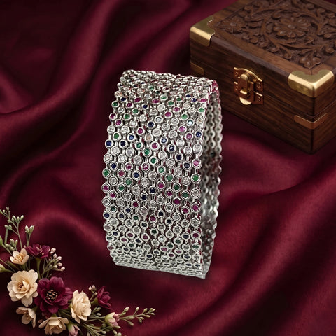 A wide silver-toned cuff bracelet covered in rows of small round gemstones in green, red, blue, and clear, displayed on draped burgundy fabric with a carved wooden box and flowers in the background.