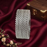 A wide silver-toned cuff bracelet covered in rows of clear rhinestones, displayed on draped burgundy satin fabric with a wooden jewelry box and flowers in the background.