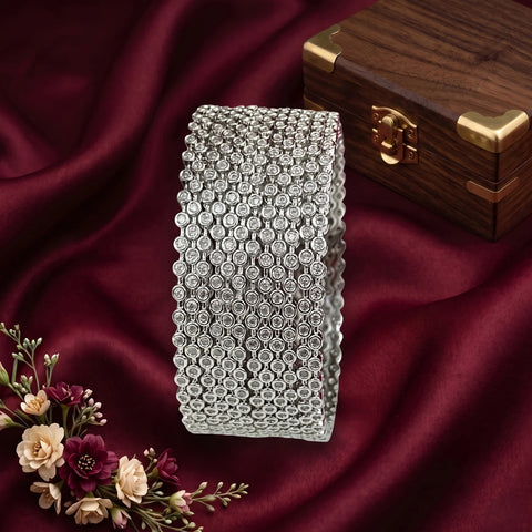 A wide silver-toned cuff bracelet covered in rows of clear rhinestones, displayed on draped burgundy satin fabric with a wooden jewelry box and flowers in the background.