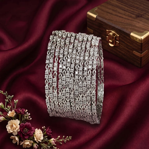 A stack of wide silver-toned bangles with intricate openwork filigree and pavé-style detailing, displayed on draped burgundy satin fabric beside a wooden jewelry box and small flowers.