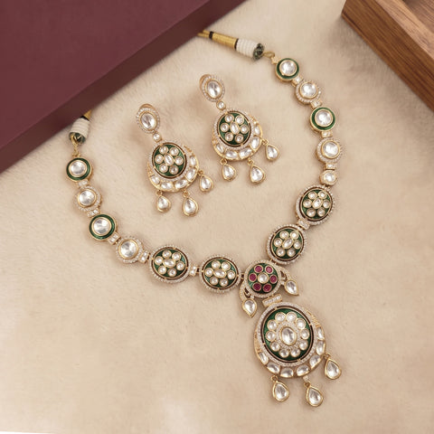 Gold-toned traditional necklace and matching earrings with green enamel, white pearl-like stones, and a small red accent, arranged on a beige surface with decorative box corners.