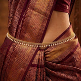 A close-up of a model wearing a traditional Indian saree in deep maroon and gold, accessorized with a gold chain waist belt decorated with small white pearls.