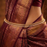 A close-up of a woman wearing a traditional Indian saree in deep maroon and gold, accessorized with a gold chain waist chain decorated with small white pearls.