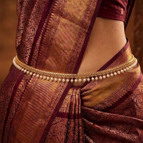 A close-up of a woman wearing a traditional Indian saree in deep maroon and gold, accessorized with a gold chain waist chain decorated with small white pearls.