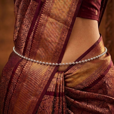 A close-up of a person wearing a metallic silver beaded waist chain (kara) over a richly patterned maroon and gold saree.