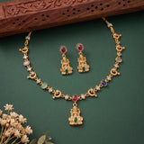 Gold-toned jewelry set with a necklace and matching earrings featuring a central deity pendant, crescent moon motifs, and small multicolored stone accents, arranged on a dark green background with a carved wooden prop and dried flowers.