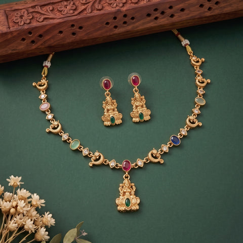 Gold-toned jewelry set with a necklace and matching earrings featuring a central deity pendant, crescent moon motifs, and small multicolored stone accents, arranged on a dark green background with a carved wooden prop and dried flowers.