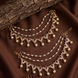 A pair of gold-toned traditional Indian chain necklaces (Kara) decorated with white faux pearls, clear rhinestones, and small red gemstone accents, arranged on a brown fabric background with a rustic wood branch.