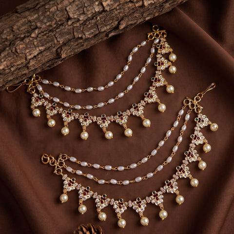 A pair of gold-toned traditional Indian chain necklaces (Kara) decorated with white faux pearls, clear rhinestones, and small red gemstone accents, arranged on a brown fabric background with a rustic wood branch.