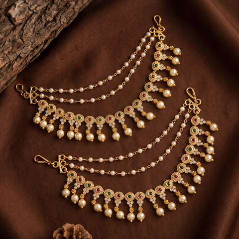 Gold-toned traditional Indian jhumka earrings with white faux pearls and colorful green, red, and pink enamel details, displayed on a dark brown fabric background with a piece of wood.