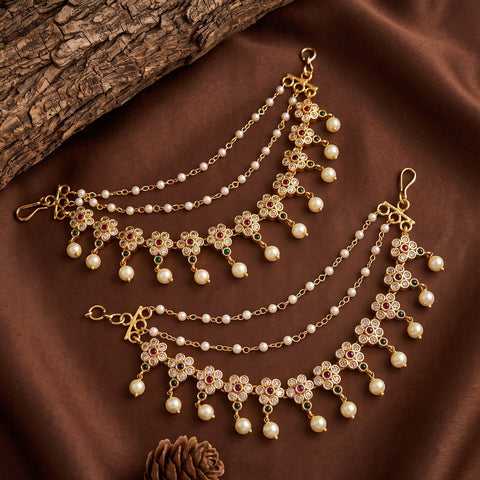 Gold-plated chain choker with floral motifs, white pearls, and small red and green stone accents, displayed on a brown satin fabric with a wood slice and pinecone prop.