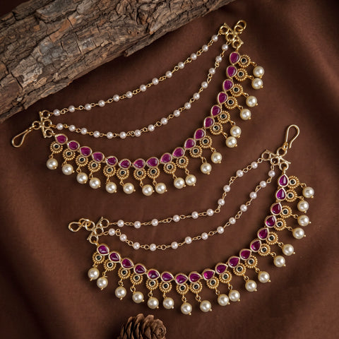 Gold-toned traditional Indian jhumka chains decorated with white faux pearls and pink gemstones, arranged on a brown fabric background with a piece of wood.