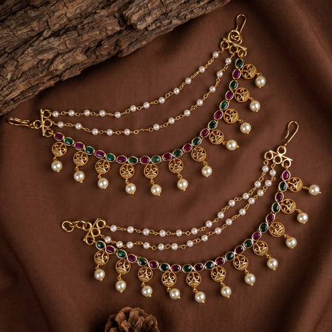 Gold-toned traditional Indian jhumka chain earrings with white faux pearls, green and red stones, and dangling ornate motifs, arranged on a brown fabric background with a piece of wood.