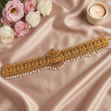 An ornate gold-toned traditional waist chain or choker with intricate floral embroidery, a central decorative motif, and dangling pearl-like beads, arranged on draped blush satin fabric with pink and white roses and a lit candle nearby.