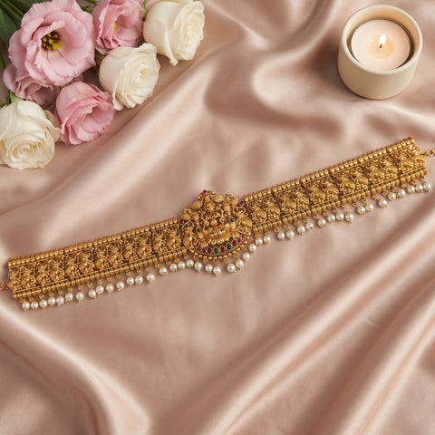 An ornate gold-toned traditional waist chain or choker with intricate floral embroidery, a central decorative motif, and dangling pearl-like beads, arranged on draped blush satin fabric with pink and white roses and a lit candle nearby.