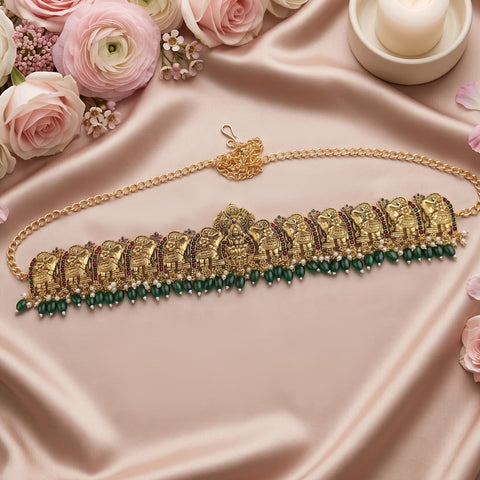 An ornate gold-toned traditional necklace with elephant motifs and a row of dark green bead drops, arranged on pink satin fabric with roses and a candle in the background.