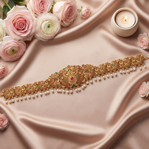 An ornate gold traditional Indian choker necklace with intricate filigree work, small green and pink stones, and dangling white pearls, displayed on draped champagne satin fabric with pink roses and a lit candle.