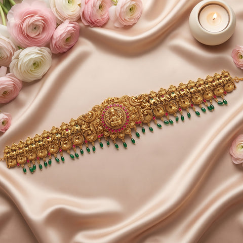 An ornate gold-toned traditional jewelry piece with a central medallion, floral detailing, and dangling green and pink accents, arranged on draped satin fabric with pink and white flowers and a lit candle.