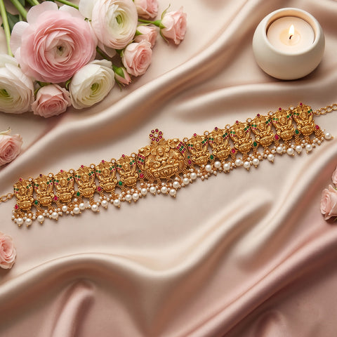 An ornate gold traditional Indian choker necklace with intricate motifs, red and green stone accents, and a row of white pearls, displayed on draped pink satin fabric with pink and white roses and a lit candle.