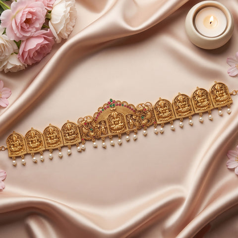 An ornate gold-toned traditional necklace with multiple deity motifs, hanging pearl beads, and small red and green stone accents, arranged on draped satin fabric with flowers and a lit candle in the background.
