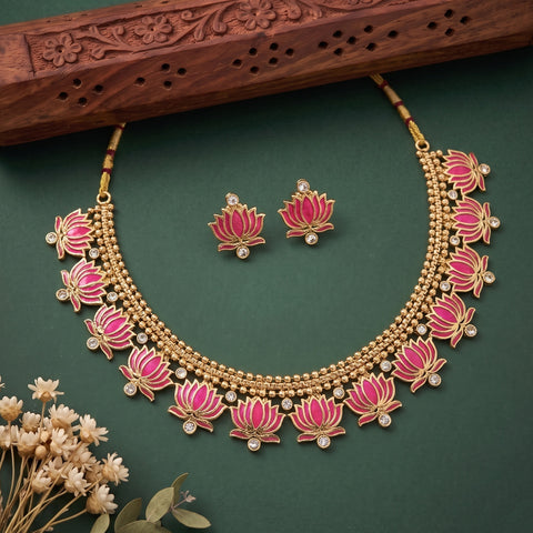 Gold-toned necklace and matching earrings with pink lotus flower motifs and small clear stone accents, arranged on a dark green background with decorative dried flowers and a carved wooden prop.