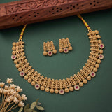 Gold-toned traditional necklace and matching earrings with red gemstone accents, arranged on a dark green background with a carved wooden prop and small dried flowers.
