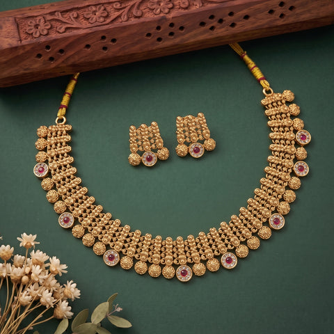 Gold-toned traditional necklace and matching earrings with red gemstone accents, arranged on a dark green background with a carved wooden prop and small dried flowers.